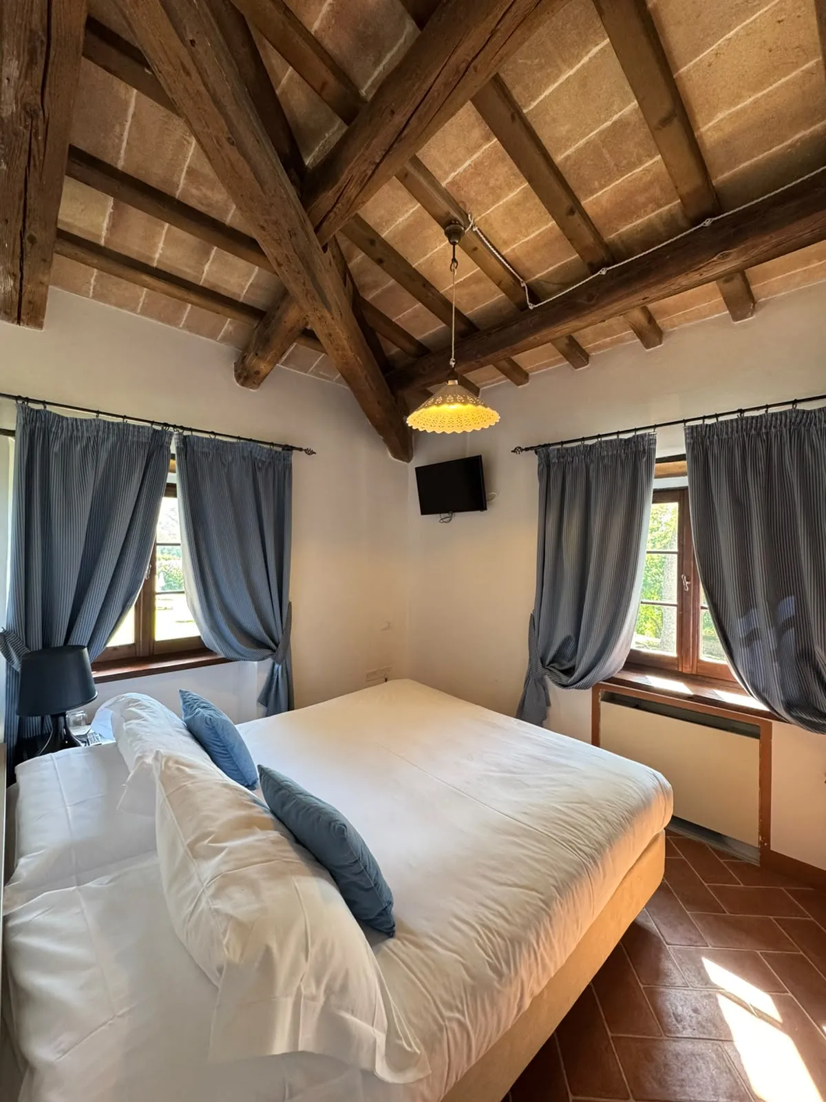 Camera Standard con letto matrimoniale, tende azzurre, soffitto a travi con lampada e pavimento in cotto