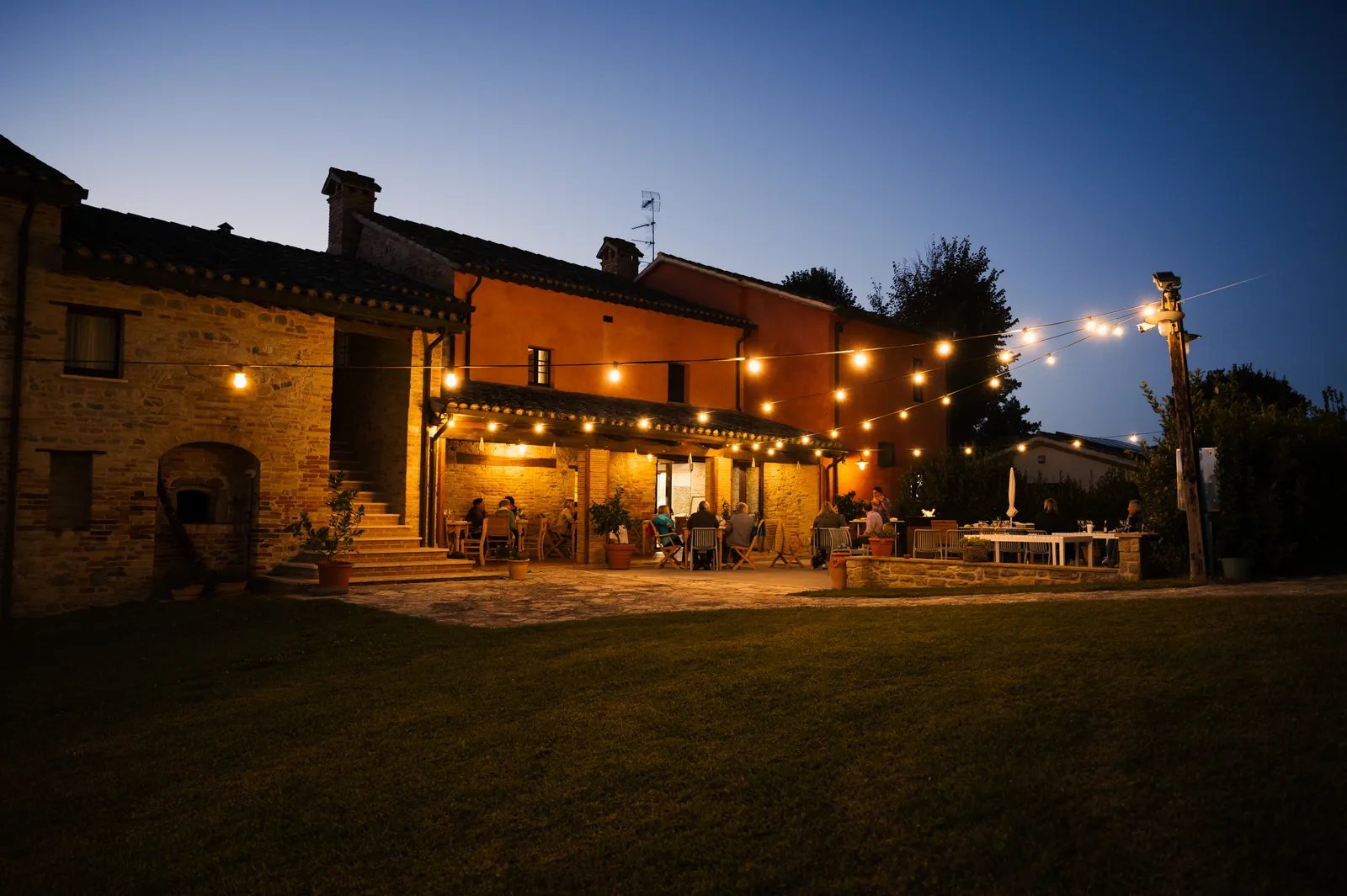 Il porticato e il cortile di Ca' Virginia illuminati per una cena serale, con le luci a stringa e gli ospiti a tavola